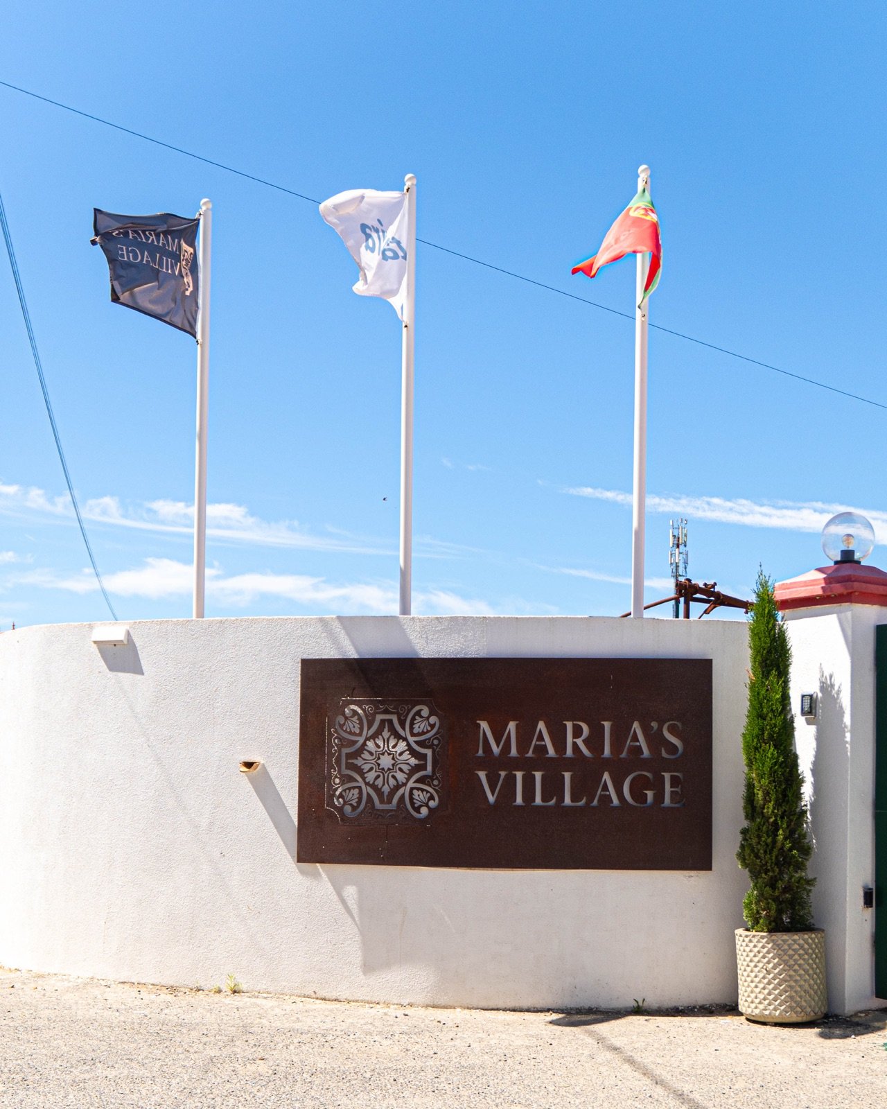 Entrada do Maria's Village com placa em muro branco e bandeiras